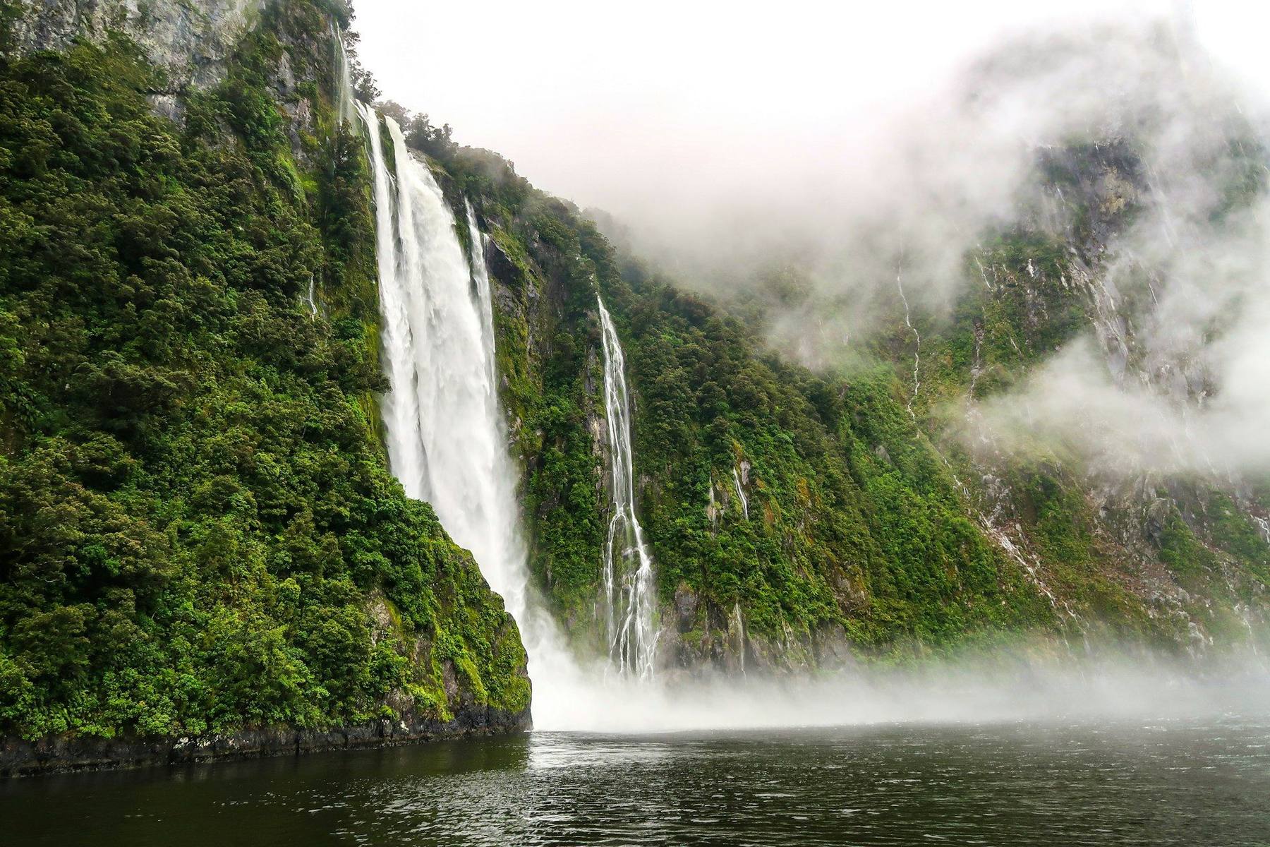 A hiker on a boardwalk in the Clinton Valley on the Milford Track, with waterfalls dropping off sheer Fiordland cliffs and beech forest on either side